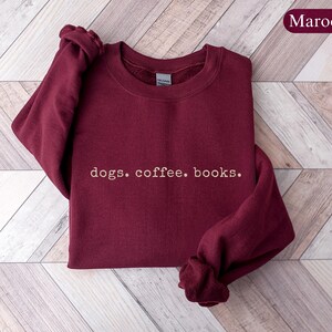 May include: Maroon sweatshirt with the words "dogs. coffee. books." in a simple, white font. The sweatshirt is folded on a light brown and white wooden surface. The word "Maroon" is in a maroon oval in the upper right corner.