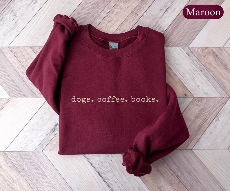 May include: Maroon sweatshirt with the words "dogs. coffee. books." in a simple, white font. The sweatshirt is folded on a light brown and white wooden surface. The word "Maroon" is in a maroon oval in the upper right corner.