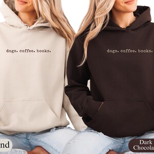 May include: Two hooded sweatshirts, one sand-colored and one dark chocolate, with the text "dogs. coffee. books." printed in a simple font. Both sweatshirts have a front pocket and a drawstring hood. The models are wearing blue jeans.