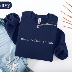 May include: Navy blue sweatshirt with the embroidered text "dogs. coffee. books." A silver necklace with a small pendant is draped across the sweatshirt. The sweatshirt is folded next to a pair of blue jeans.