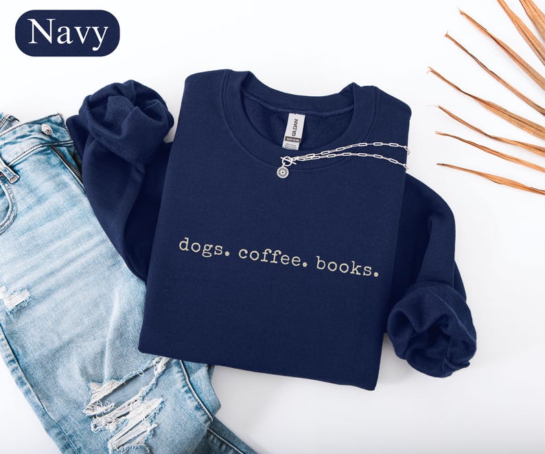 May include: Navy blue sweatshirt with the embroidered text "dogs. coffee. books." A silver necklace with a small pendant is draped across the sweatshirt. The sweatshirt is folded next to a pair of blue jeans.
