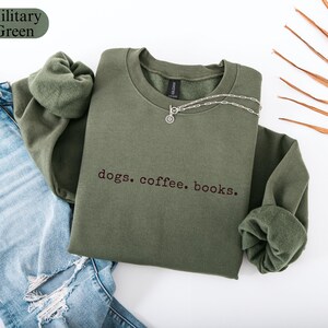May include: A military green sweatshirt with the embroidered text "dogs. coffee. books." A silver necklace with a circular pendant rests on the shirt. The sweatshirt is paired with blue jeans.