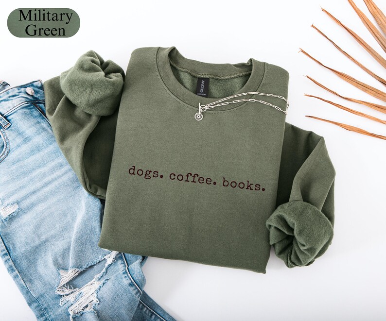 May include: A military green sweatshirt with the embroidered text "dogs. coffee. books." A silver necklace with a circular pendant rests on the shirt. The sweatshirt is paired with blue jeans.