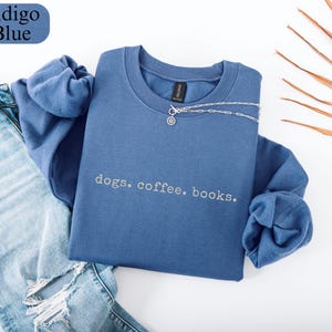 May include: An indigo blue sweatshirt with the words "dogs. coffee. books." in a light grey font. A silver necklace with a small pendant is draped over the sweatshirt. The sweatshirt is next to a pair of blue jeans.