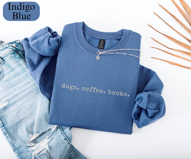 May include: An indigo blue sweatshirt with the words "dogs. coffee. books." in a light grey font. A silver necklace with a small pendant is draped over the sweatshirt. The sweatshirt is next to a pair of blue jeans.