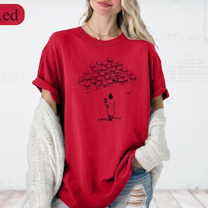 Op de afbeelding: Rood T-shirt met een zwart-wit illustratie van een persoon en een kudde schapen. Het shirt heeft korte mouwen en een ronde halslijn. Het woord "Red" staat in een rode ovaal linksboven.