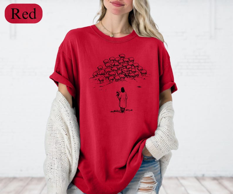 Op de afbeelding: Rood T-shirt met een zwart-wit illustratie van een persoon en een kudde schapen. Het shirt heeft korte mouwen en een ronde halslijn. Het woord "Red" staat in een rode ovaal linksboven.