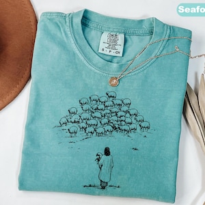 Op de afbeelding: Zeegroen t-shirt met een ronde hals en korte mouwen. Het shirt heeft een zwarte afbeelding van een herder met een schaap en een kudde schapen. Een goudkleurige ketting met een hanger is over het shirt gedrapeerd.