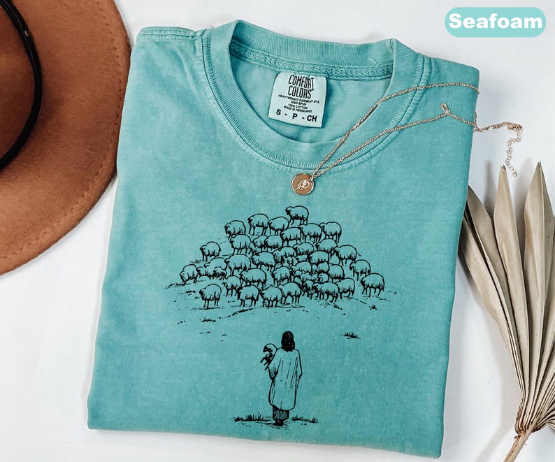 Op de afbeelding: Zeegroen t-shirt met een ronde hals en korte mouwen. Het shirt heeft een zwarte afbeelding van een herder met een schaap en een kudde schapen. Een goudkleurige ketting met een hanger is over het shirt gedrapeerd.