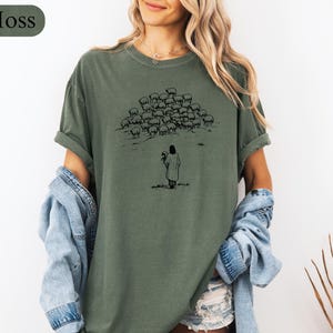 Op de afbeelding: Mosgroen t-shirt met een zwarte illustratie van een persoon die bloemen vasthoudt, met een kudde schapen op de achtergrond. Het shirt heeft opgerolde mouwen en een relaxte pasvorm. Het woord "Moss" staat in een groene ovaal linksboven.