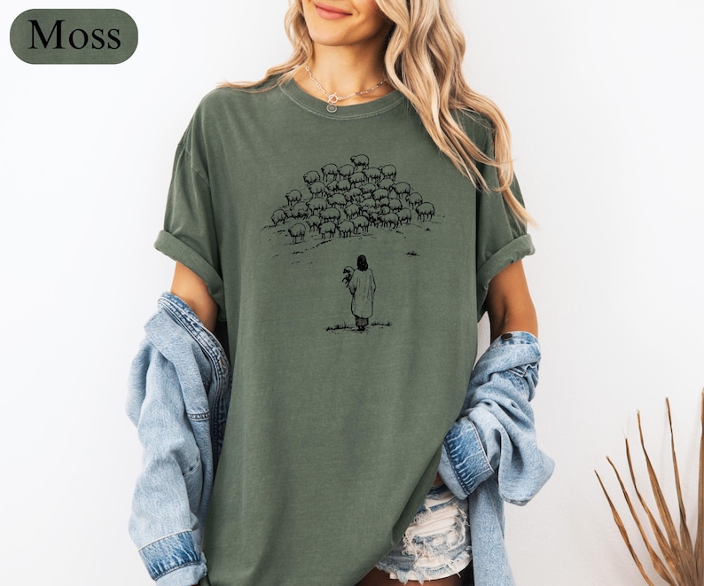 Op de afbeelding: Mosgroen t-shirt met een zwarte illustratie van een persoon die bloemen vasthoudt, met een kudde schapen op de achtergrond. Het shirt heeft opgerolde mouwen en een relaxte pasvorm. Het woord "Moss" staat in een groene ovaal linksboven.