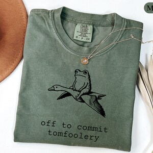 May include: A moss green crew neck t-shirt featuring a black graphic of a frog riding a flying goose. Below the graphic, the text reads off to commit tomfoolery. The shirt has a Comfort Colors brand label inside the collar indicating size S.