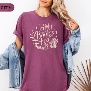 May include: Berry-colored t-shirt with the text "In My Bookish Era" in a circular design with mushroom and leaf illustrations. The shirt has short sleeves and a relaxed fit. A denim jacket is worn over the shirt.