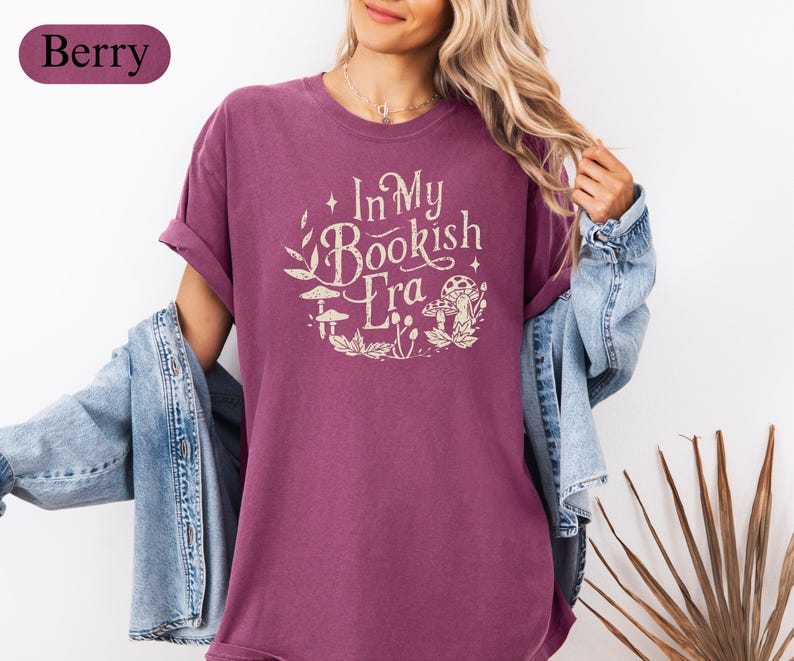May include: Berry-colored t-shirt with the text "In My Bookish Era" in a circular design with mushroom and leaf illustrations. The shirt has short sleeves and a relaxed fit. A denim jacket is worn over the shirt.