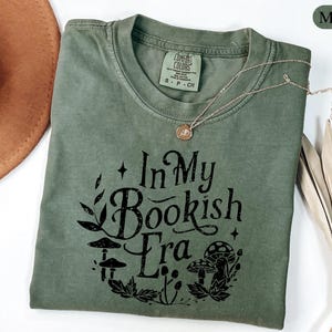 May include: A moss green t-shirt with the text "In My Bookish Era" in black, featuring mushroom and leaf graphics. The shirt has a crew neck and short sleeves. A gold necklace is draped across the shirt.