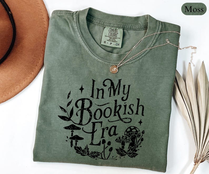 May include: A moss green t-shirt with the text "In My Bookish Era" in black, featuring mushroom and leaf graphics. The shirt has a crew neck and short sleeves. A gold necklace is draped across the shirt.