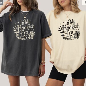 May include: Two oversized t-shirts, one pepper-colored and one ivory, are displayed. Each shirt features the text "In My Bookish Era" in a whimsical font, surrounded by botanical illustrations. The shirts are casual and comfortable.