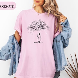 Op de afbeelding: Lichtroze t-shirt met een zwart-witte illustratie van een persoon met schapen. Het woord "Blossom" staat linksboven gedrukt. Het shirt heeft een losse pasvorm.