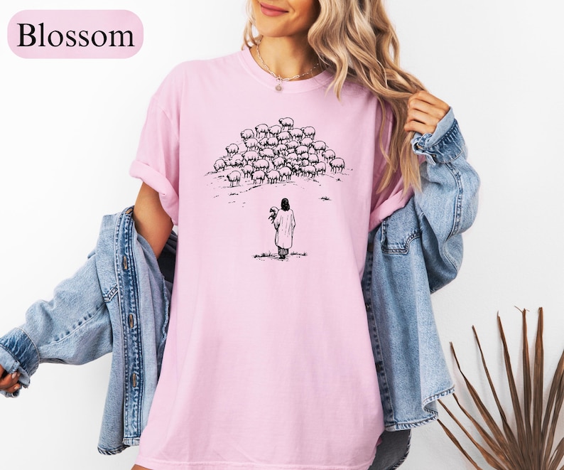 Op de afbeelding: Lichtroze t-shirt met een zwart-witte illustratie van een persoon met schapen. Het woord "Blossom" staat linksboven gedrukt. Het shirt heeft een losse pasvorm.