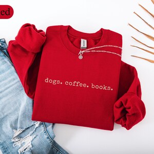 May include: A red sweatshirt with the words "dogs. coffee. books." in white script. The sweatshirt is paired with blue jeans and a silver necklace. The word "Red" is in a red oval in the upper left corner.