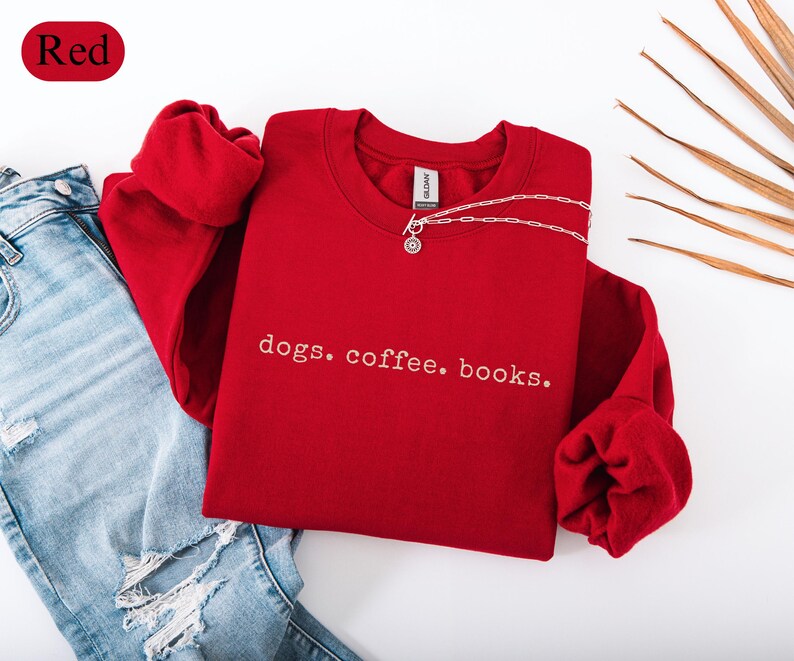 May include: A red sweatshirt with the words "dogs. coffee. books." in white script. The sweatshirt is paired with blue jeans and a silver necklace. The word "Red" is in a red oval in the upper left corner.