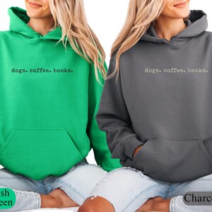 May include: Two hooded sweatshirts, one Irish green and one charcoal gray, are displayed. Each sweatshirt has a front pocket and the text "dogs. coffee. books." printed across the chest. The sweatshirts are shown on a person.