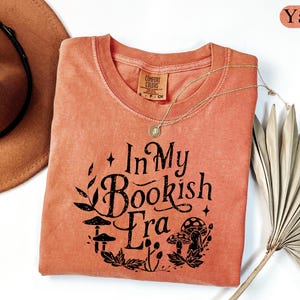 May include: A yam-colored t-shirt with the text "In My Bookish Era" in black, featuring a botanical design with mushrooms and leaves. A gold necklace and a brown hat are also visible.