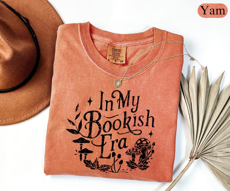 May include: A yam-colored t-shirt with the text "In My Bookish Era" in black, featuring a botanical design with mushrooms and leaves. A gold necklace and a brown hat are also visible.