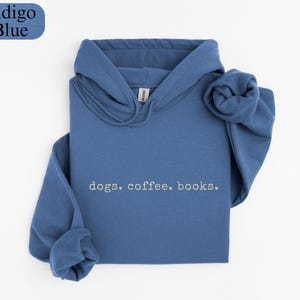 May include: Indigo blue hoodie with the words "dogs. coffee. books." in a simple font. The hoodie is folded neatly, with the hood and sleeves visible. The top left corner has a blue oval with the words "Indigo Blue".