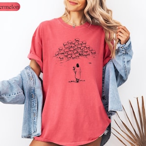 Op de afbeelding: Een watermeloenkleurig T-shirt met een zwart-wit illustratie van een persoon die bloemen vasthoudt, tegenover een kudde schapen. Het shirt heeft een relaxte pasvorm en korte mouwen. Het woord "Watermelon" staat in een roze ovaal linksboven.