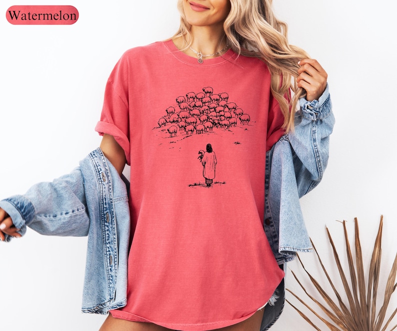 Op de afbeelding: Een watermeloenkleurig T-shirt met een zwart-wit illustratie van een persoon die bloemen vasthoudt, tegenover een kudde schapen. Het shirt heeft een relaxte pasvorm en korte mouwen. Het woord "Watermelon" staat in een roze ovaal linksboven.
