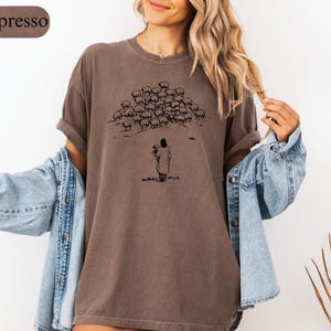 Op de afbeelding: Espresso-kleurig T-shirt met een zwarte illustratie van een herder en schapen. De herder staat in een veld en houdt een lam vast. Het shirt heeft een relaxte pasvorm en korte mouwen. Het woord "Espresso" staat in een bruin ovaal.