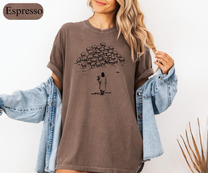 Op de afbeelding: Espresso-kleurig T-shirt met een zwarte illustratie van een herder en schapen. De herder staat in een veld en houdt een lam vast. Het shirt heeft een relaxte pasvorm en korte mouwen. Het woord "Espresso" staat in een bruin ovaal.