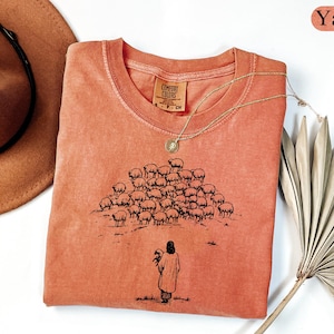 Op de afbeelding: Een koraalkleurig T-shirt met een ronde hals en een afbeelding van een herder met schapen. Het shirt heeft een Comfort Colors-label en is versierd met een gouden ketting. Een bruine hoed en gedroogde palmbladeren zijn ook in de afbeelding.