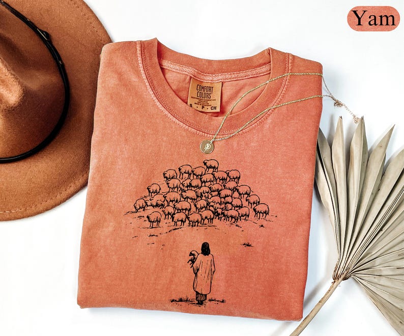 Op de afbeelding: Een koraalkleurig T-shirt met een ronde hals en een afbeelding van een herder met schapen. Het shirt heeft een Comfort Colors-label en is versierd met een gouden ketting. Een bruine hoed en gedroogde palmbladeren zijn ook in de afbeelding.