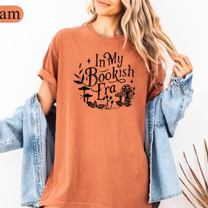 May include: A yam-colored t-shirt with the text "In My Bookish Era" in black, surrounded by a floral and mushroom design. The shirt has short sleeves and a relaxed fit.