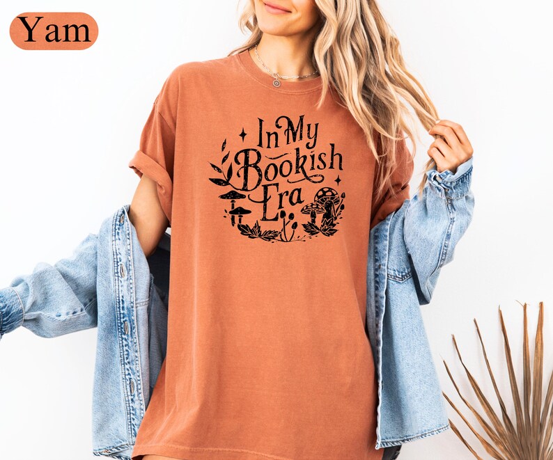 May include: A yam-colored t-shirt with the text "In My Bookish Era" in black, surrounded by a floral and mushroom design. The shirt has short sleeves and a relaxed fit.