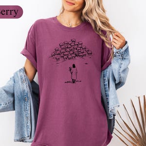 Op de afbeelding: Bessenkleurig T-shirt met een zwarte illustratie van een figuur met bloemen, tegenover een kudde schapen. Het shirt heeft korte mouwen en een relaxte pasvorm. Het woord "Berry" staat in een bordeauxrood ovaal linksboven.