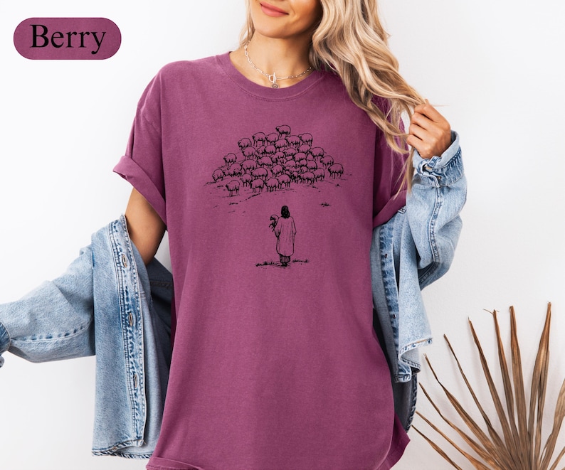 Op de afbeelding: Bessenkleurig T-shirt met een zwarte illustratie van een figuur met bloemen, tegenover een kudde schapen. Het shirt heeft korte mouwen en een relaxte pasvorm. Het woord "Berry" staat in een bordeauxrood ovaal linksboven.