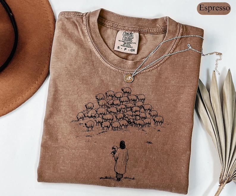 Op de afbeelding: Espresso-kleurig Comfort Colors T-shirt met een ronde hals. Het shirt heeft een afbeelding van een persoon die een schaap vasthoudt, met een kudde schapen op de achtergrond. Een zilveren ketting met een hanger ligt op het shirt.