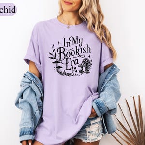 May include: A light purple t-shirt with the text "In My Bookish Era" in a black, whimsical font, surrounded by mushroom and leaf illustrations. The shirt is part of a casual outfit, paired with a denim jacket.