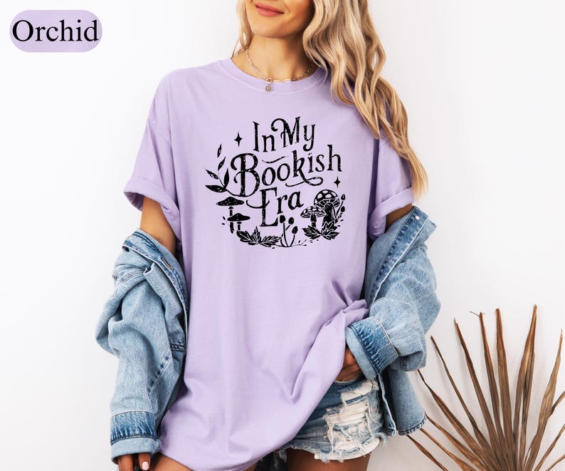 May include: A light purple t-shirt with the text "In My Bookish Era" in a black, whimsical font, surrounded by mushroom and leaf illustrations. The shirt is part of a casual outfit, paired with a denim jacket.