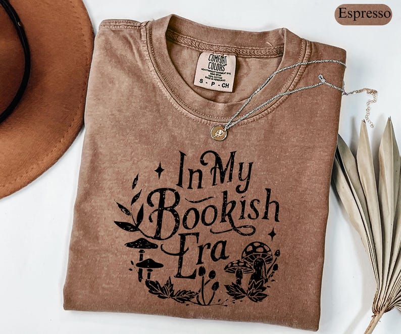 May include: Espresso-colored t-shirt with the text "In My Bookish Era" in a whimsical font, featuring mushroom and leaf illustrations. A silver necklace with a pendant rests on the shirt. The shirt is a Comfort Colors brand.