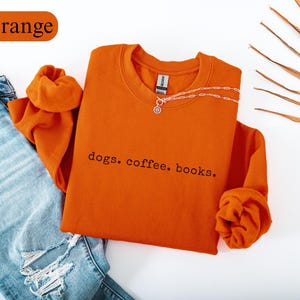 May include: An orange crewneck sweatshirt with the text "dogs. coffee. books." in black. The sweatshirt is paired with light blue ripped jeans and a silver necklace. The word "Orange" is in a rectangular shape in the top left corner.