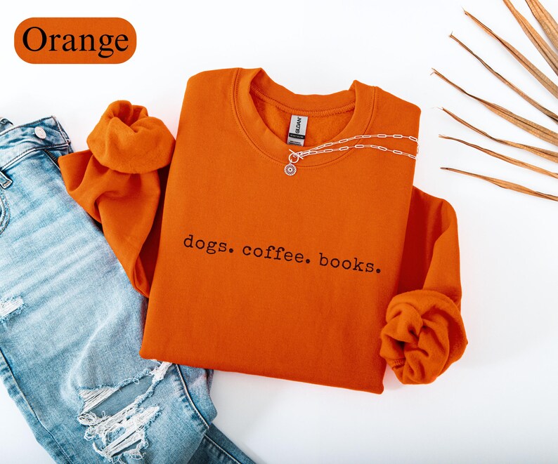 May include: An orange crewneck sweatshirt with the text "dogs. coffee. books." in black. The sweatshirt is paired with light blue ripped jeans and a silver necklace. The word "Orange" is in a rectangular shape in the top left corner.