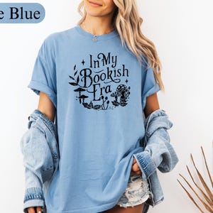 May include: Ice blue t-shirt with rolled sleeves. The shirt features the text "In My Bookish Era" in a black, circular design with mushrooms and leaves. The model is wearing a denim jacket over the shirt.