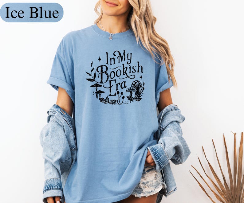 May include: Ice blue t-shirt with rolled sleeves. The shirt features the text "In My Bookish Era" in a black, circular design with mushrooms and leaves. The model is wearing a denim jacket over the shirt.