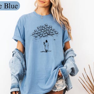 Op de afbeelding: IJsblauwe t-shirt met een zwart-wit illustratie van een persoon die een schaap vasthoudt, tegenover een kudde schapen. De tekst "Ice Blue" staat boven het shirt. Het shirt heeft opgerolde mouwen.