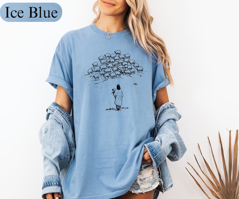 Op de afbeelding: IJsblauwe t-shirt met een zwart-wit illustratie van een persoon die een schaap vasthoudt, tegenover een kudde schapen. De tekst "Ice Blue" staat boven het shirt. Het shirt heeft opgerolde mouwen.