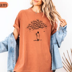 Op de afbeelding: Een terracotta-kleurig t-shirt met een zwarte illustratie van een herder en schapen. De herder houdt een lam vast en de schapen staan op een heuvel. Het woord "Yam" staat in een oranje ovaal in de linkerbovenhoek.